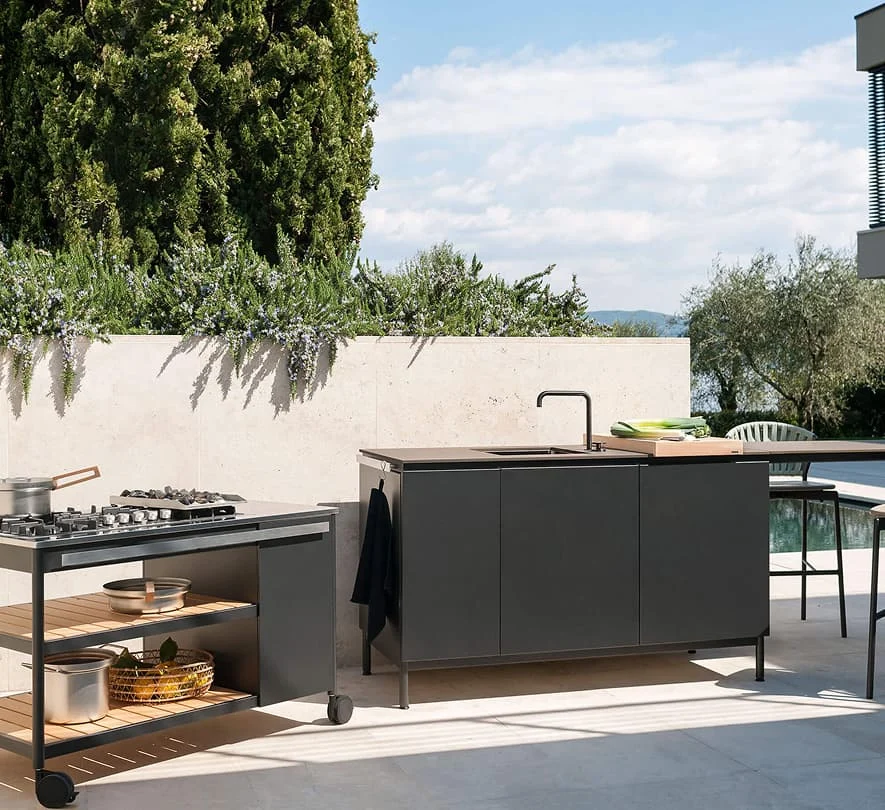 Moderne Außenküche mit schwarzem Kochmodul, Spüle und Arbeitsfläche im Garten, umgeben von mediterranen Pflanzen und Poolblick.