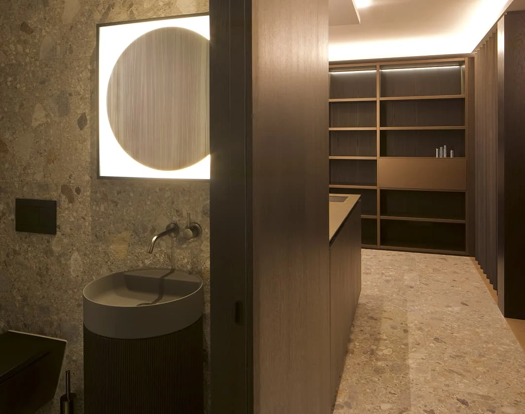 Moderner Bad und Wellnessbereich mit beleuchtetem Rundspiegel, steinverkleideter Wand, kompaktem Waschbecken und angrenzendem Raum mit großen offenen Holzregalen.