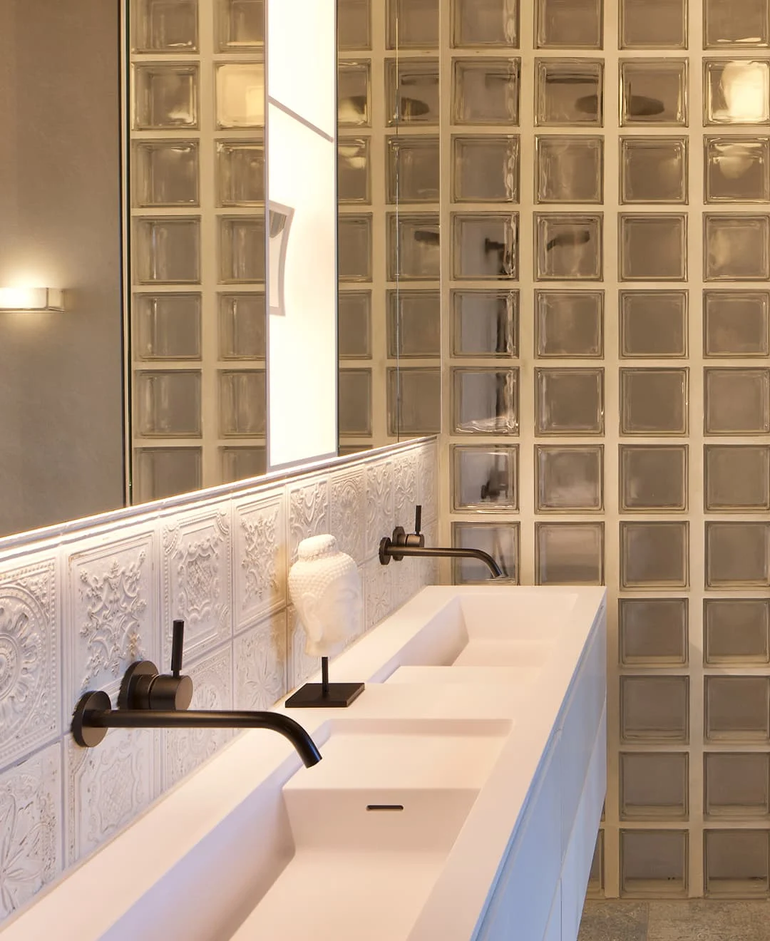 Modernes Badezimmer mit Doppelwaschbecken, mattschwarzen Armaturen, weißer Ornamentfliesen-Rückwand und Glasbausteinwand.