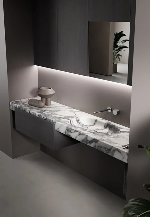 Modernes Badezimmer mit einer eleganten Waschtischplatte aus weißem Marmor mit dunkler Maserung, minimalistischer Wandarmatur, dunklen Schrankfronten und indirekter LED-Beleuchtung für ein hochwertiges Interior-Design.