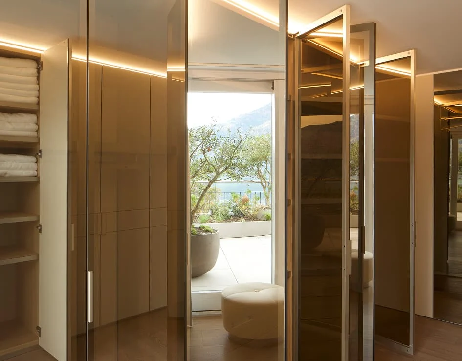 Begehbarer Kleiderschrank mit Glasfronten und Blick auf Terrasse mit Seelandschaft.