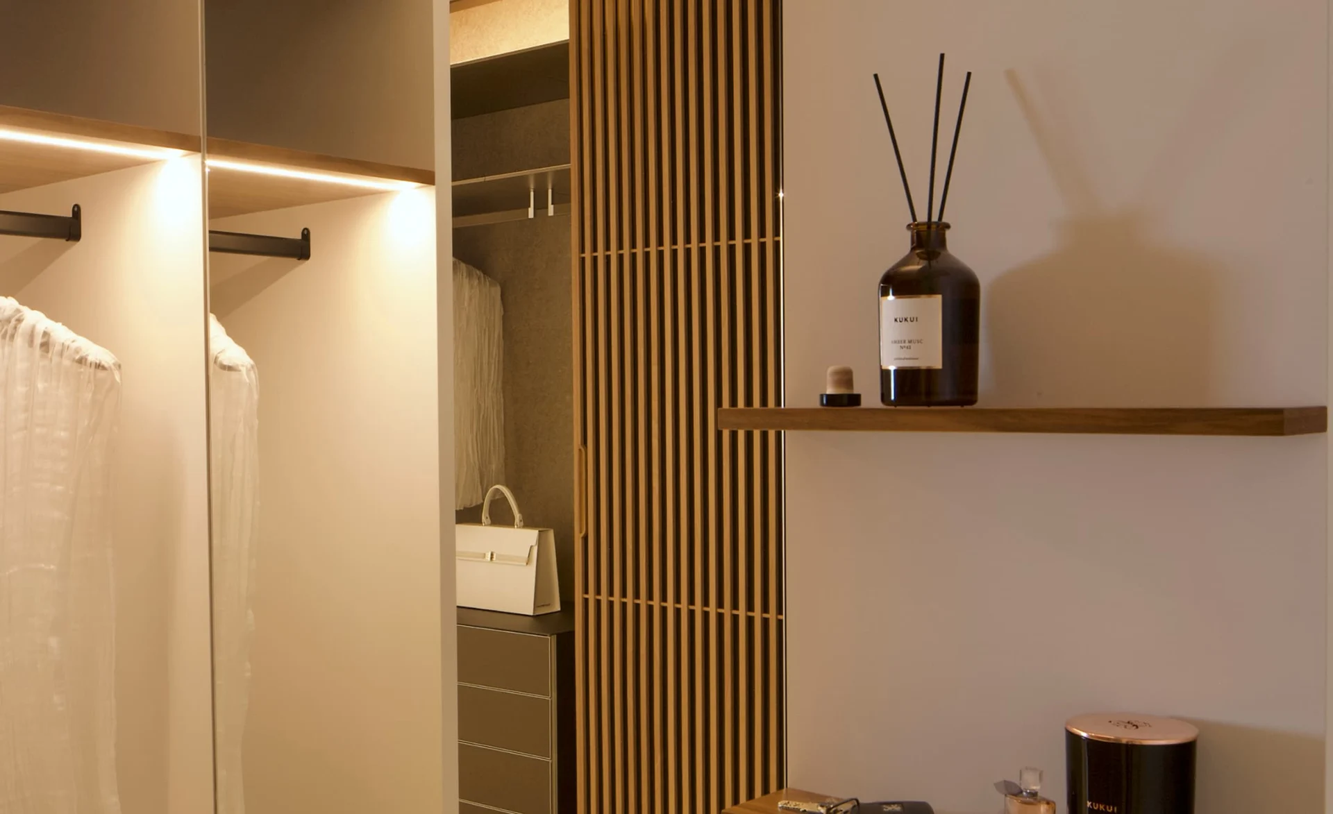 Moderner begehbarer Kleiderschrank mit Holzlamellen, offener Kleiderstange und dekorativer Duftflasche auf Wandregal.