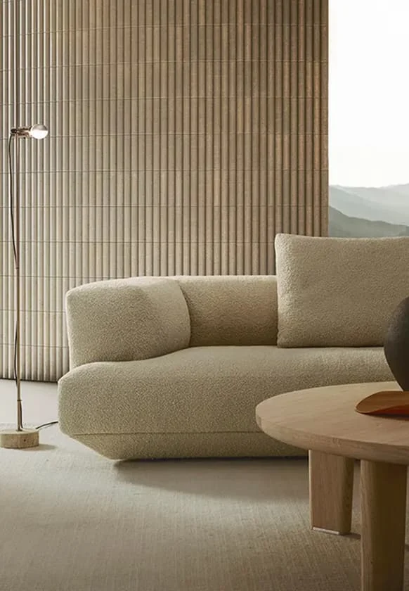Beiges Designersofa aus Bouclé-Stoff vor gerippter Wandverkleidung mit minimalistischer Stehleuchte und rundem Holztisch im modernen Wohnraum.