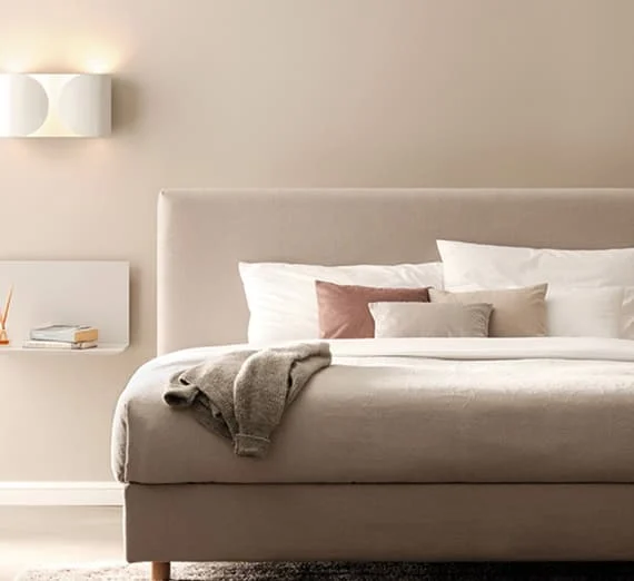 Modernes, beigefarbenes Polsterbett mit mehreren Kissen und einer abgelegten Strickjacke in einem hellen, minimalistischen Schlafzimmer.