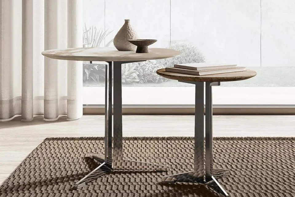 Zwei moderne Beistelltische mit runden Platten aus Stein und Holz, schlanken Metallgestellen und dekorativen Objekten im Vordergrund, arrangiert auf einem strukturierten Teppich vor einer großen Fensterfront.