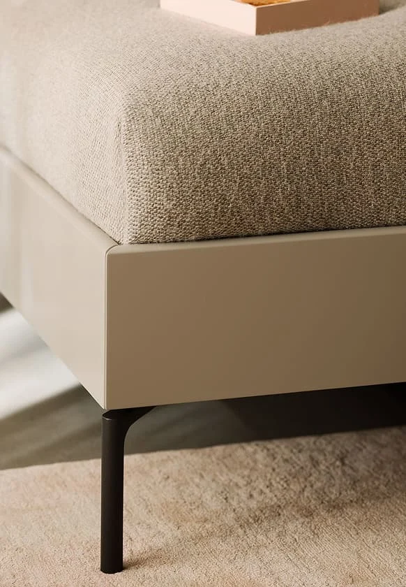 Detailaufnahme eines modernen Bettgestells in Beige mit gepolsterter Matratzenkante und filigranem schwarzen Metallfuß.