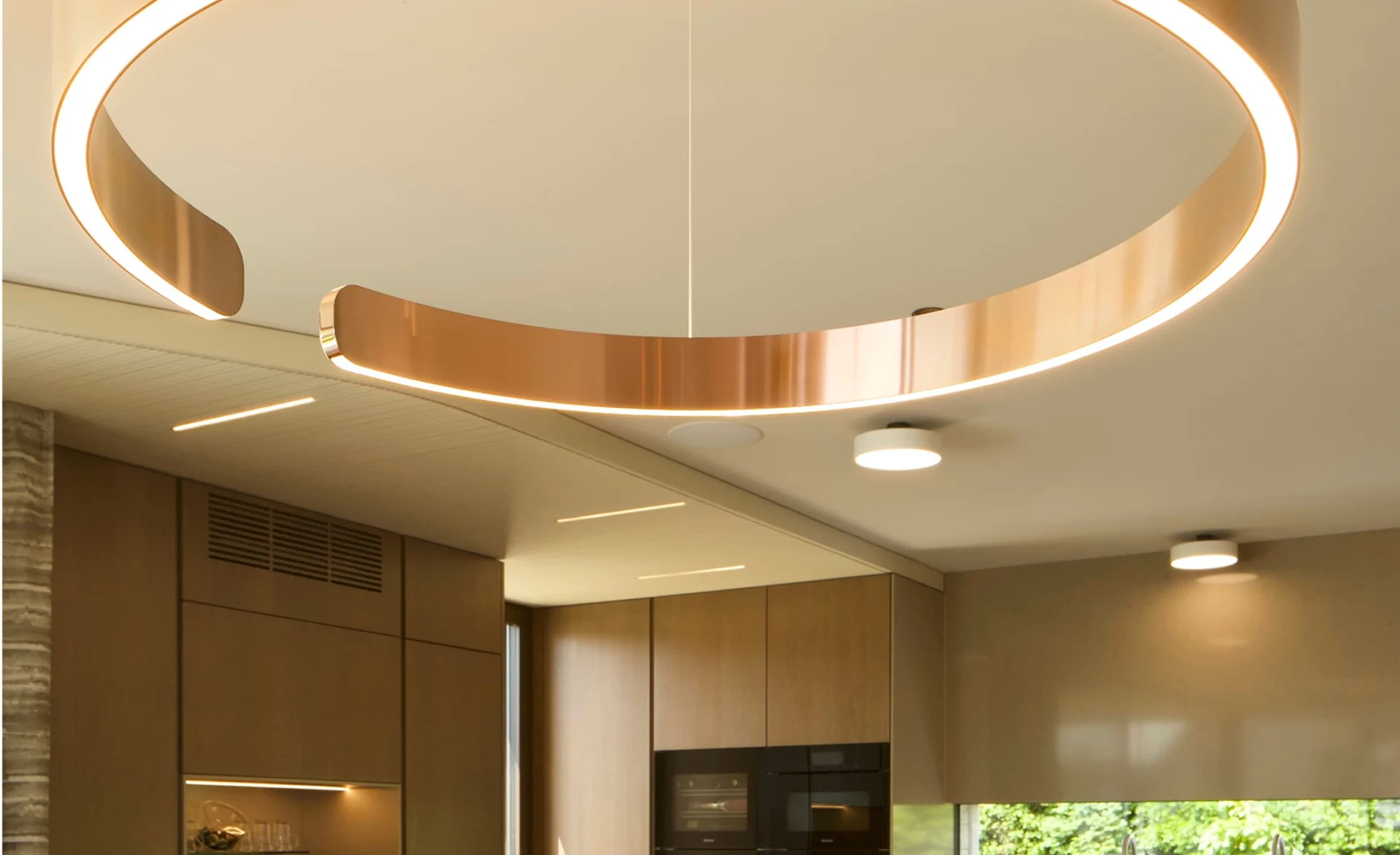 Große ringförmige Deckenleuchte mit warmem LED Licht über einer modernen Küche mit Holzfronten.