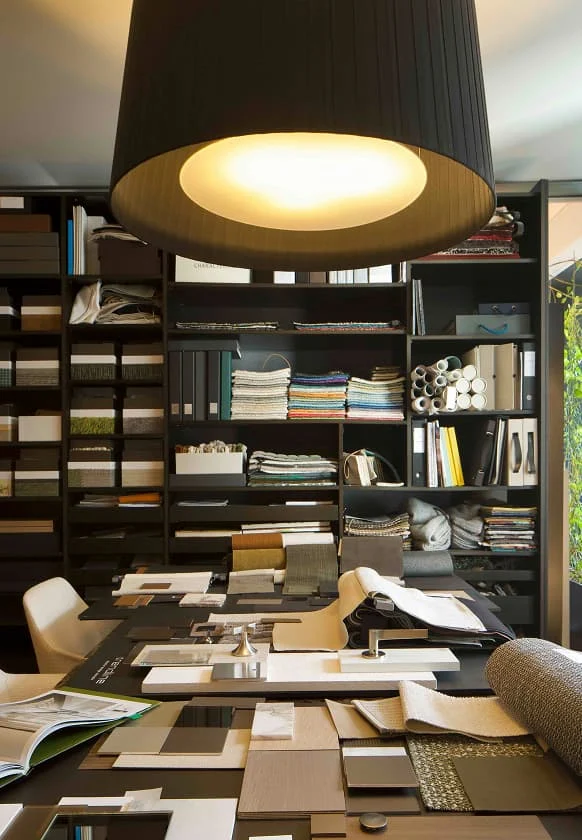 Mustertisch mit Stoffproben, Materialmustern und Katalogen vor einem großen Regal voller Design- und Einrichtungsproben.