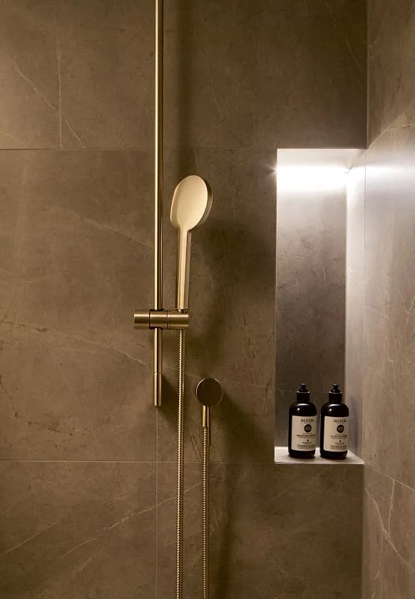 Goldene Duscharmatur vor einer grauen Steinwand mit beleuchtetem Nischenregal und Duschprodukten.
