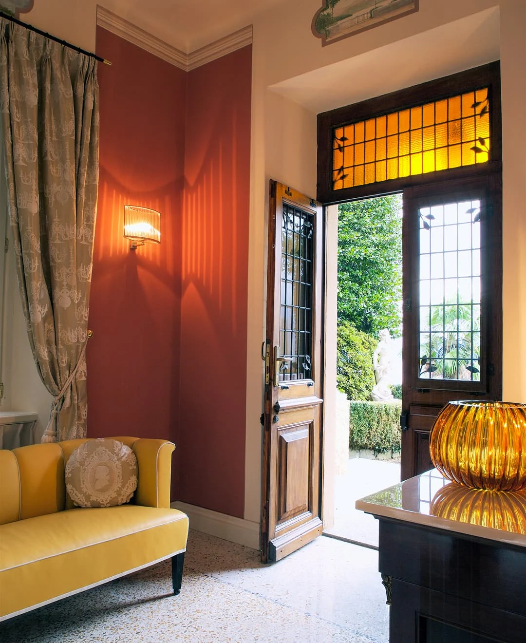 Eleganter Eingangsbereich mit gelbem Sofa, roséfarbener Wand, offener Holztür und warmem Glaslampenlicht.