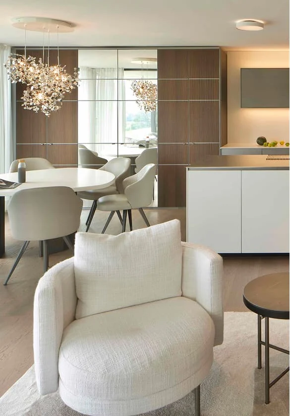 Eleganter cremefarbener Lounge-Sessel in modernem Wohn- und Essbereich mit hellem Holzboden und Designer-Leuchten.