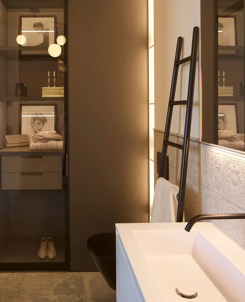 Elegantes Badezimmer mit schwarzer Leiter als Handtuchhalter, modernem Waschbecken und beleuchtetem Schrank mit dekorativen Bildern.