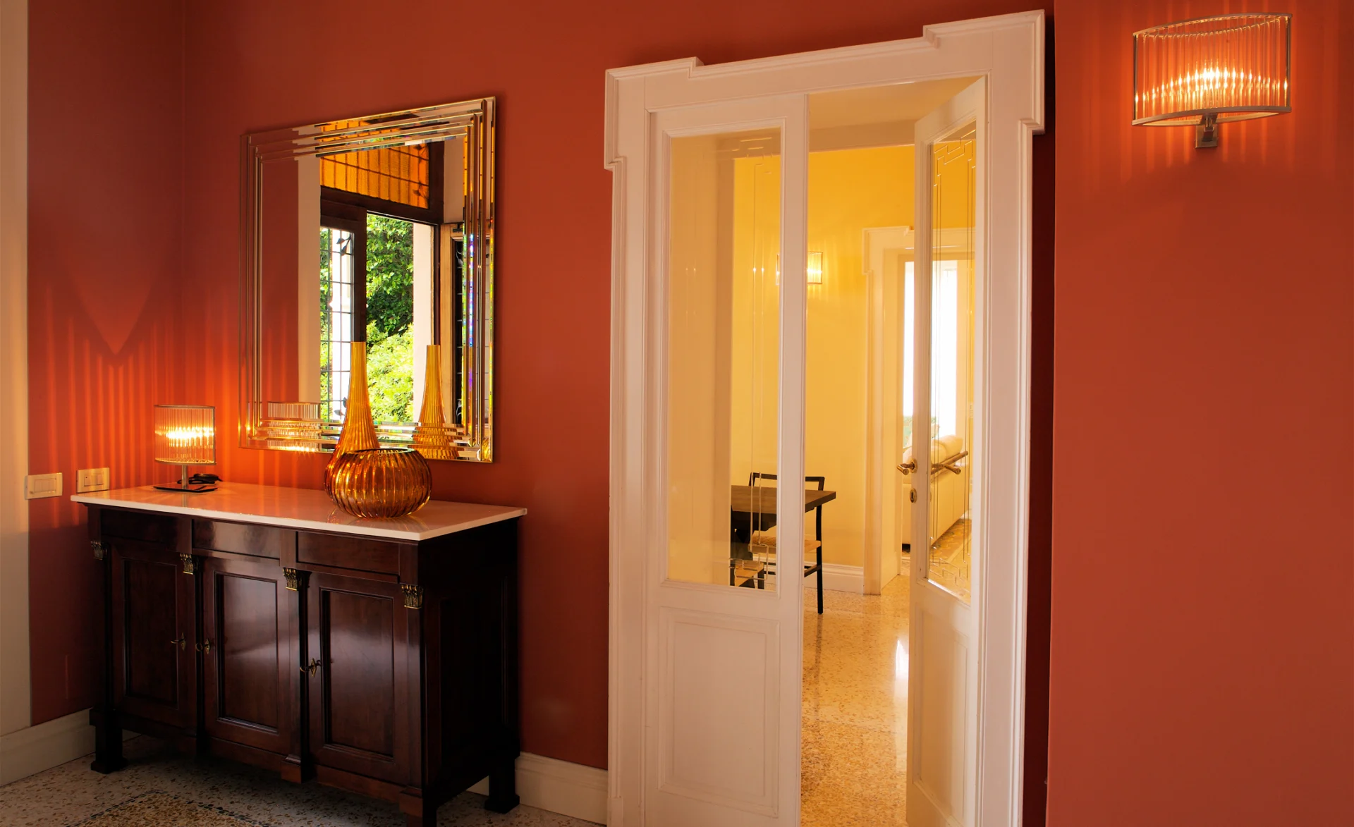 Elegantes Foyer mit dunkler Holzkommode, großem Spiegel, warmen Wandfarben und Blick in den hellen Nebenraum