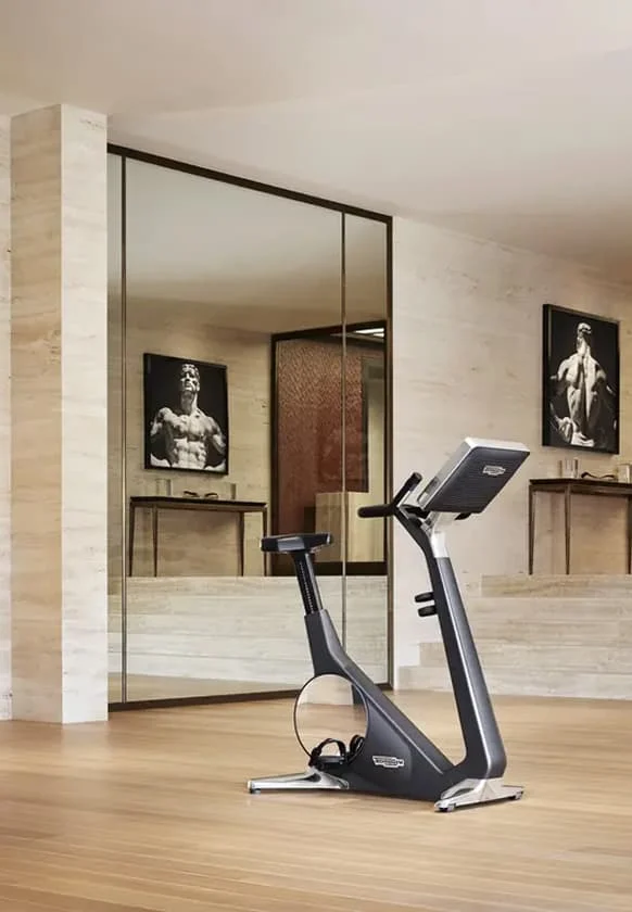 Heller Fitnessraum mit modernem Indoor-Bike auf Holzfußboden, großen Spiegel- und Glasflächen sowie Wandbildern von muskulösen Athleten.