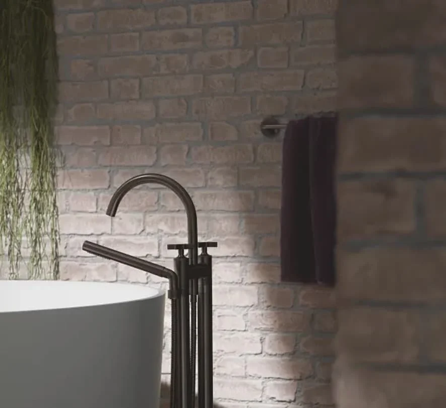 Freistehende schwarze Badewannenarmatur vor heller Ziegelwand mit hängender Pflanze und Handtuch im Hintergrund.