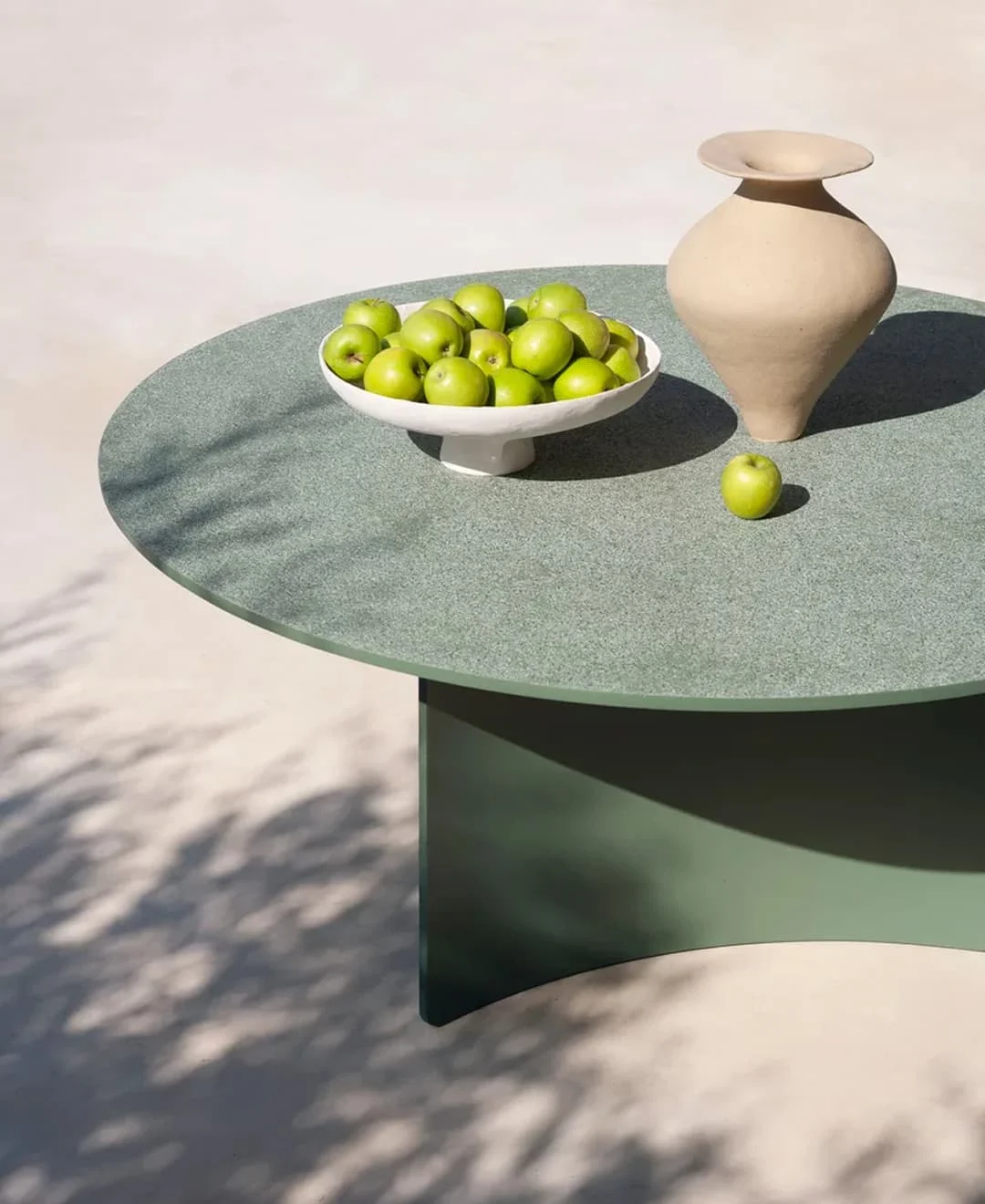 Runder Gartentisch in Grün mit einer Schale voller grüner Äpfel und einer beigen Keramikvase im Sonnenlicht.