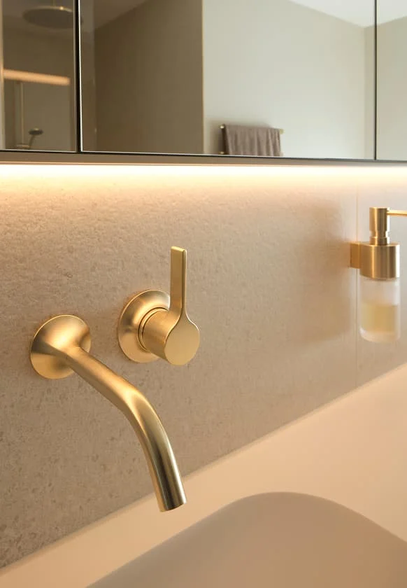 Goldfarbene Wandarmatur über einem modernen Waschbecken mit warmer LED-Beleuchtung und integriertem Seifenspender.