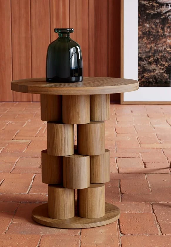 Runder Holztisch mit skulpturaler, zylindrischer Basis und dunkler Glasvase auf terrakottafarbenem Boden.