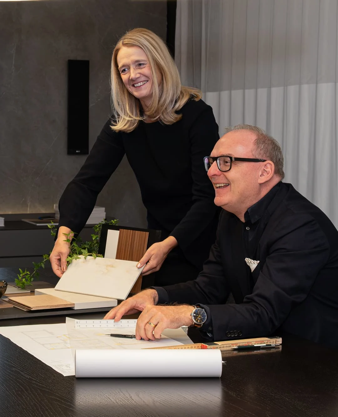 Zwei Innenarchitekten bei der Materialauswahl mit Mustern und Bauplänen am Tisch.