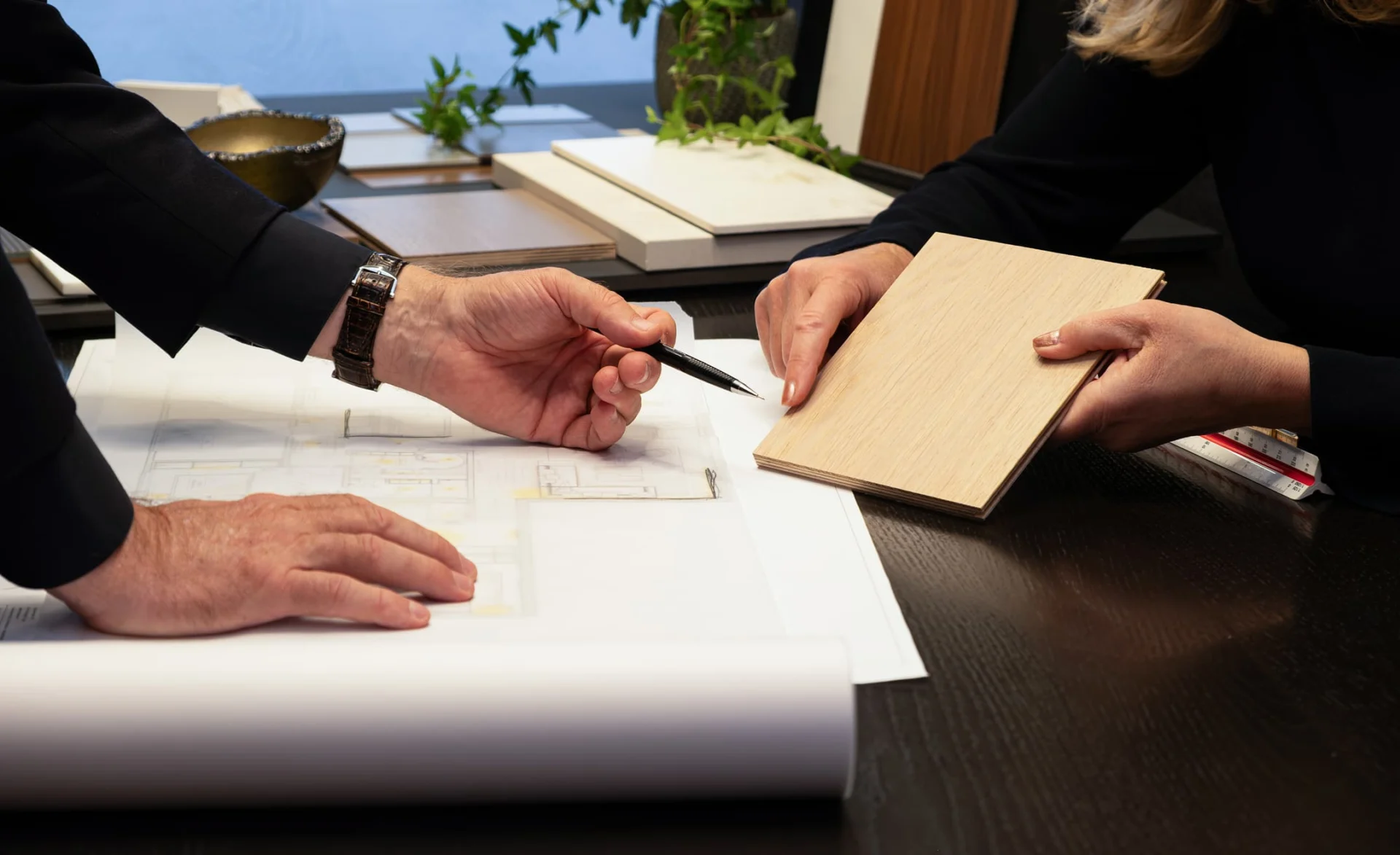 Zwei Personen besprechen Innenraumpläne und vergleichen eine helle Holzprobe auf einem Tisch mit Zeichnungen und Materialmustern.