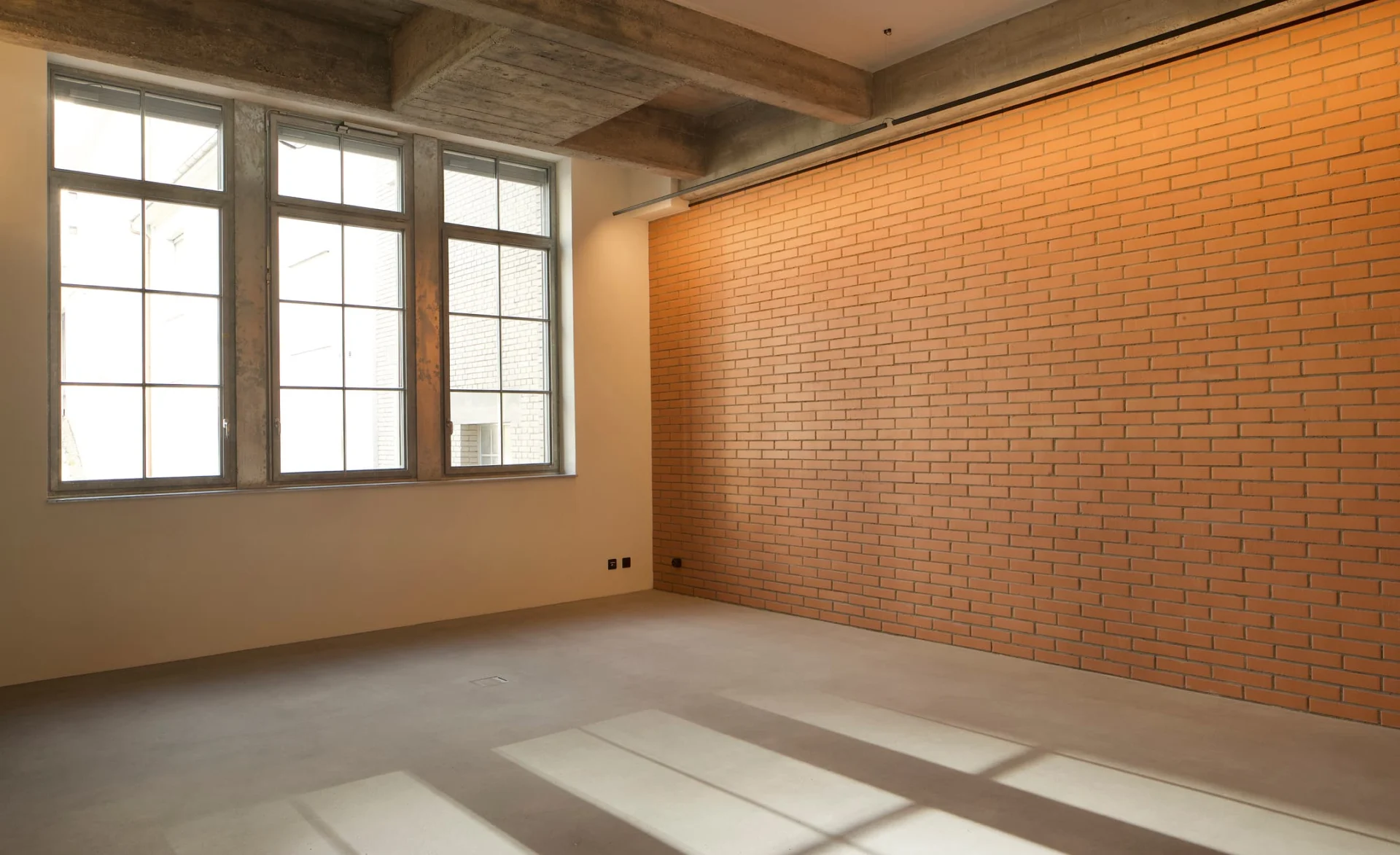 Leerer Innenraum mit großer Fensterfront, sichtbarer Betondecke und langer Ziegelwand in moderner Industriearchitektur.
