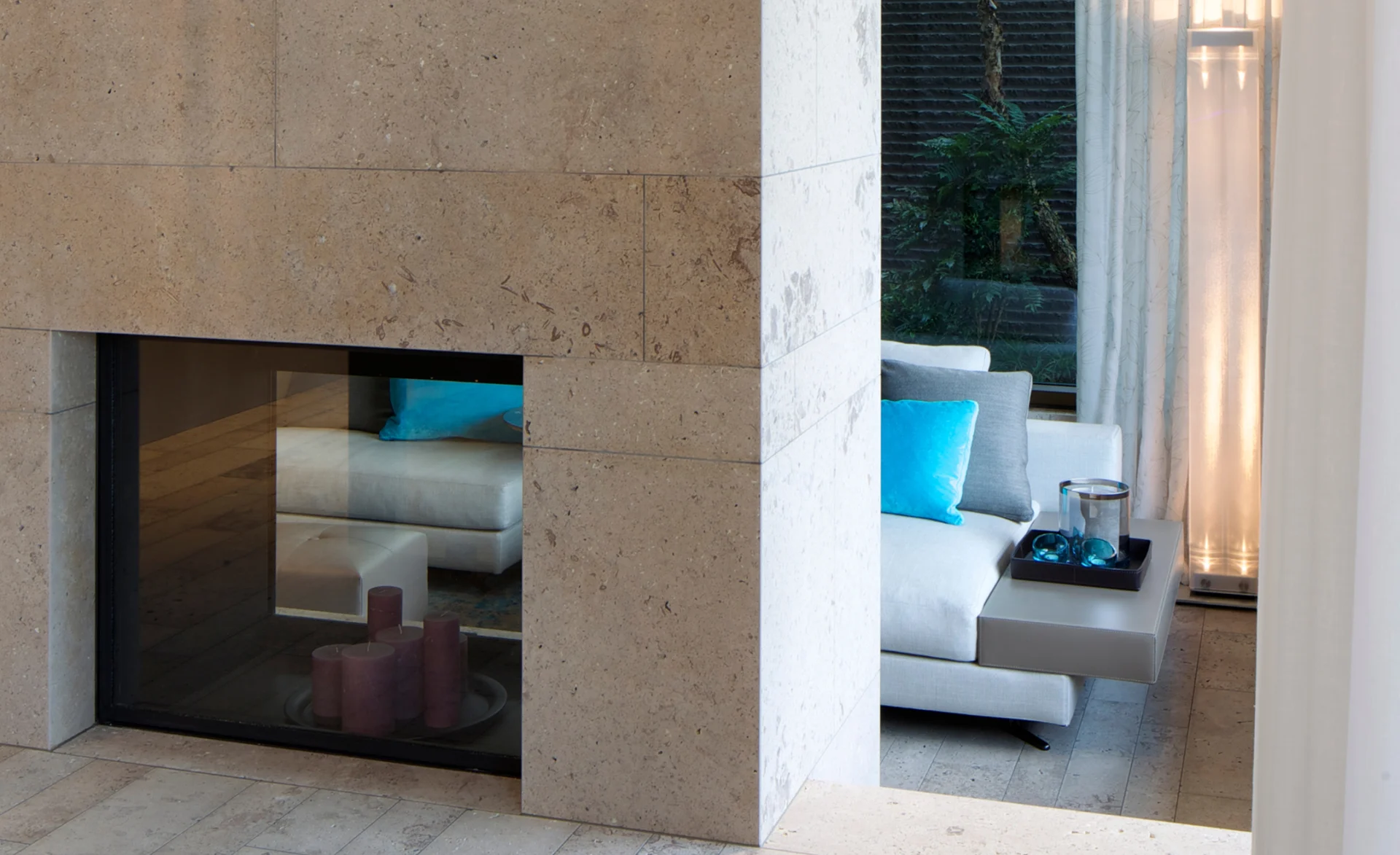 Natursteinkamin mit Blick in den Wohnbereich mit hellen Sofas und türkisfarbenen Akzenten.