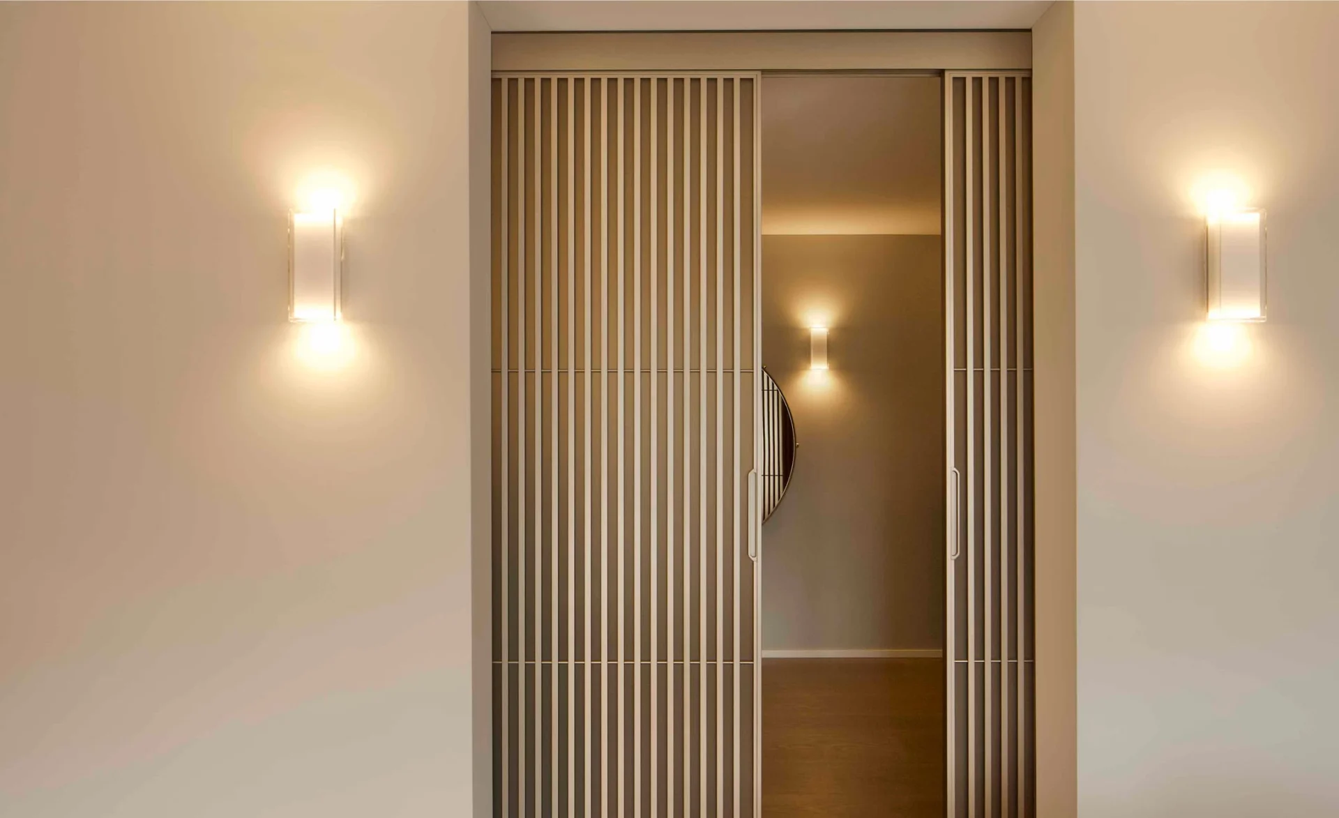 Lamellentür mit Blick in einen warm beleuchteten Flur, flankiert von Wandlampen an einer hellen Wand in moderner Innenarchitektur.