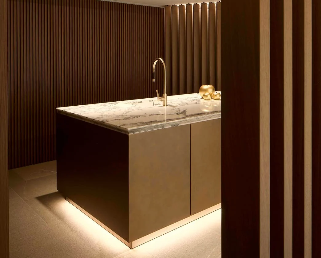 Elegante Kücheninsel mit weißer Marmorarbeitsplatte, goldener Armatur und umlaufender Holzlamellenwand im Trendline Showroom.