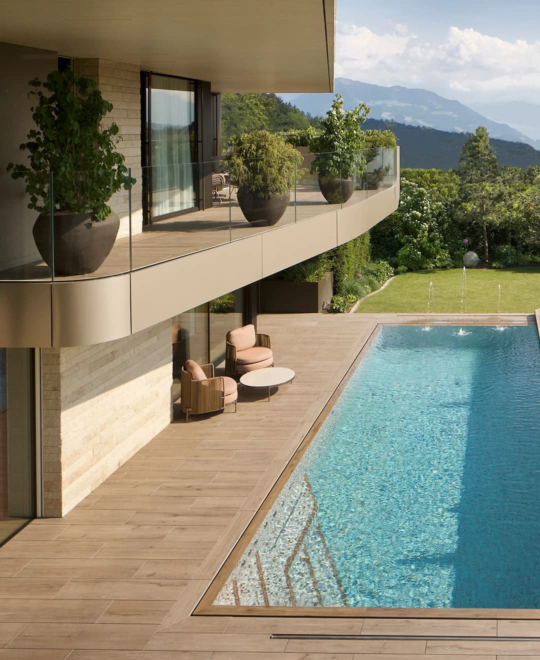 Moderner Außenbereich einer Luxusvilla mit Pool, Holzterrasse, großen Pflanzgefäßen und Blick auf die umliegenden Berge.