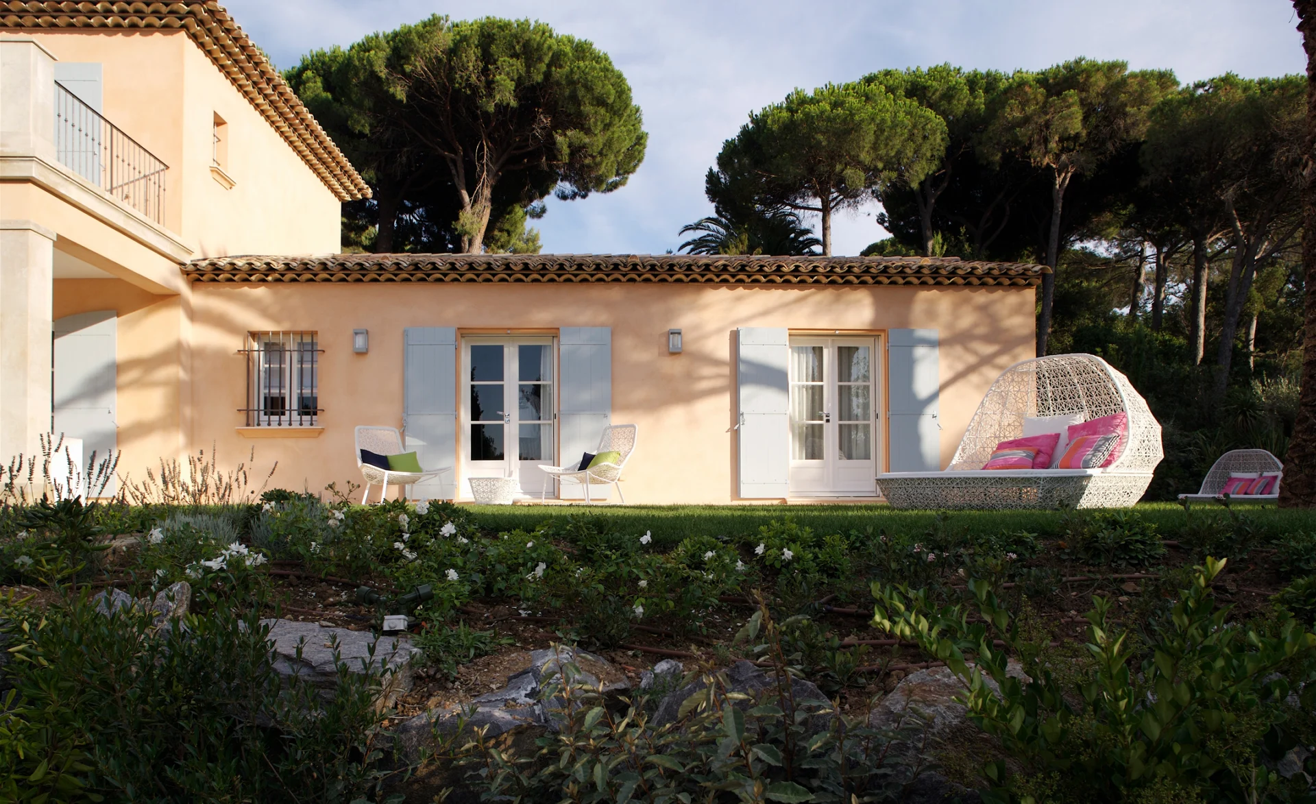 Mediterrane Villa mit hellblauen Fensterläden und Gartenlounge aus weißem Geflecht vor Pinienbäumen