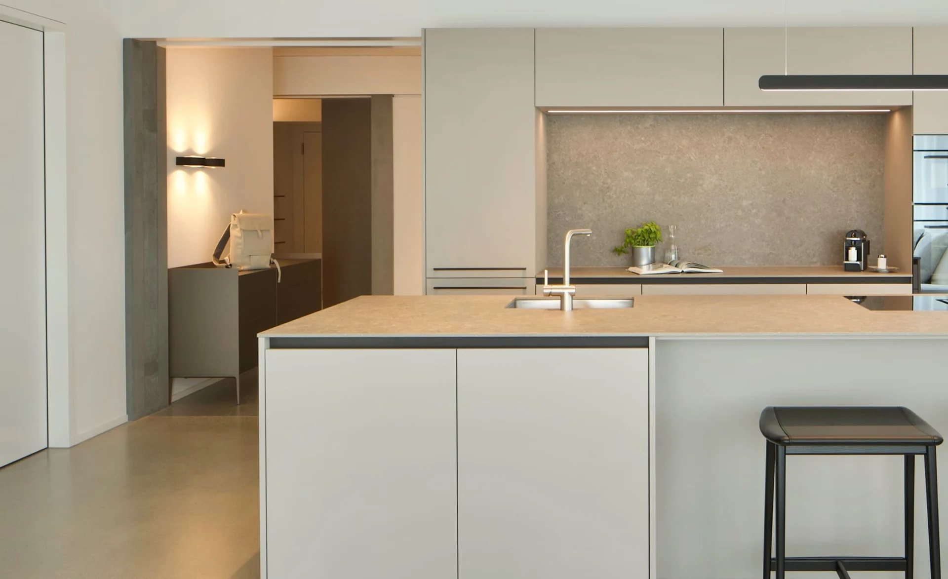 Moderne helle Küche mit großer Kochinsel, Einbauschränken in Beige und grauer Steinrückwand, dezent beleuchtet und mit Barhocker im Vordergrund.