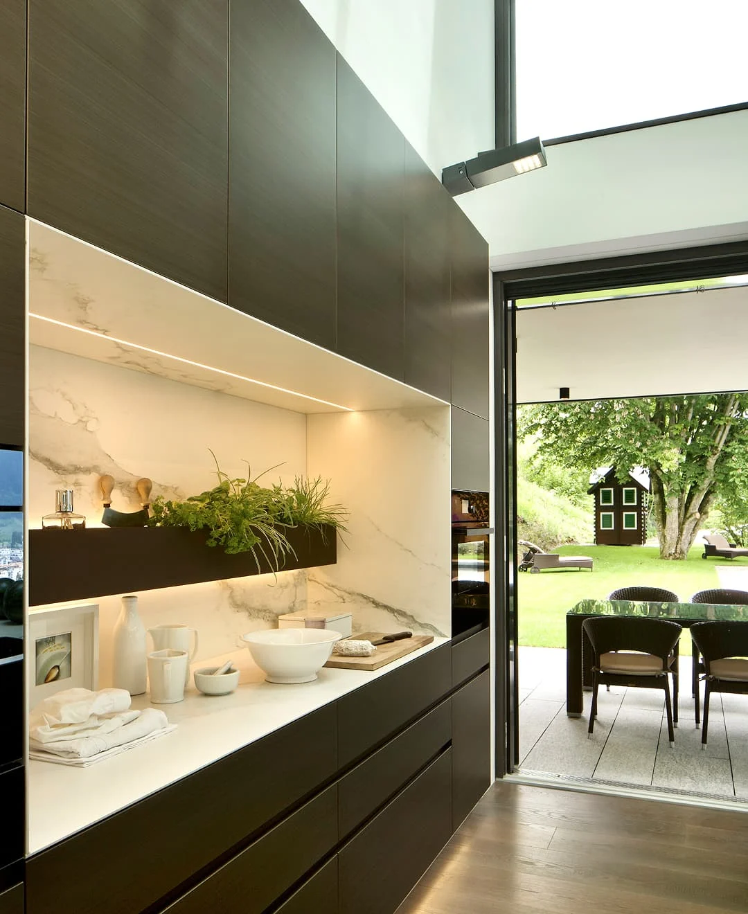 Moderne Küche mit dunklen Schränken, heller Marmorarbeitsfläche und offener Glasfront zum Garten mit Essbereich im Freien.