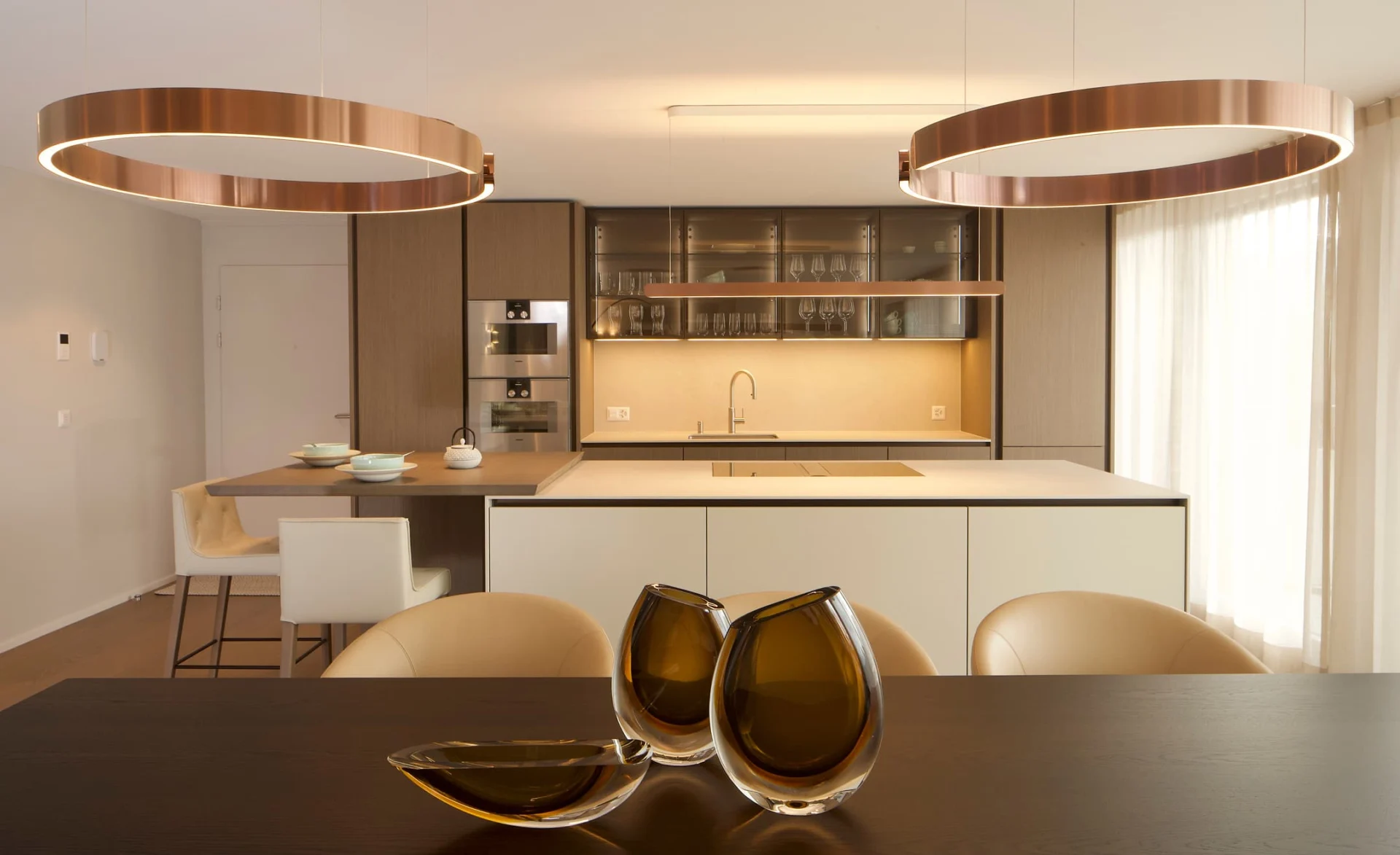 Moderne offene Küche mit großen runden Kupferleuchten, heller Kochinsel, Glasregalen und elegant gedecktem Esstisch im Vordergrund