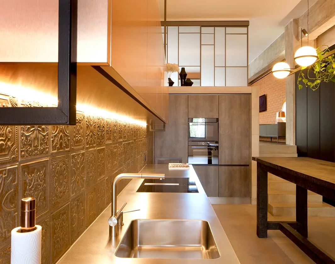 Moderne Loftküche mit metallischen Fronten, strukturierter Fliesenrückwand und hochwertiger Beleuchtung in warmem Industrial Ambiente.