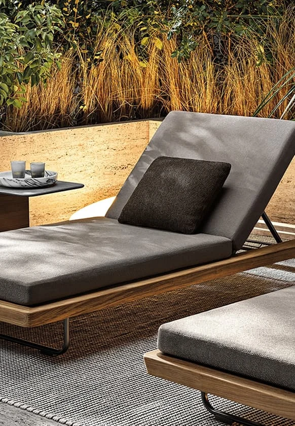 Moderne Outdoor-Liege mit grauen Polstern und Holzrahmen, daneben kleiner Beistelltisch und stimmungsvolle Gartenbeleuchtung.