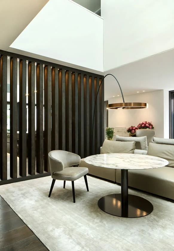 Elegante moderne Sitzecke mit rundem Marmortisch, beigem Sofa, Designerstuhl und geschwungener Stehleuchte vor dunkler Holzlamellenwand.