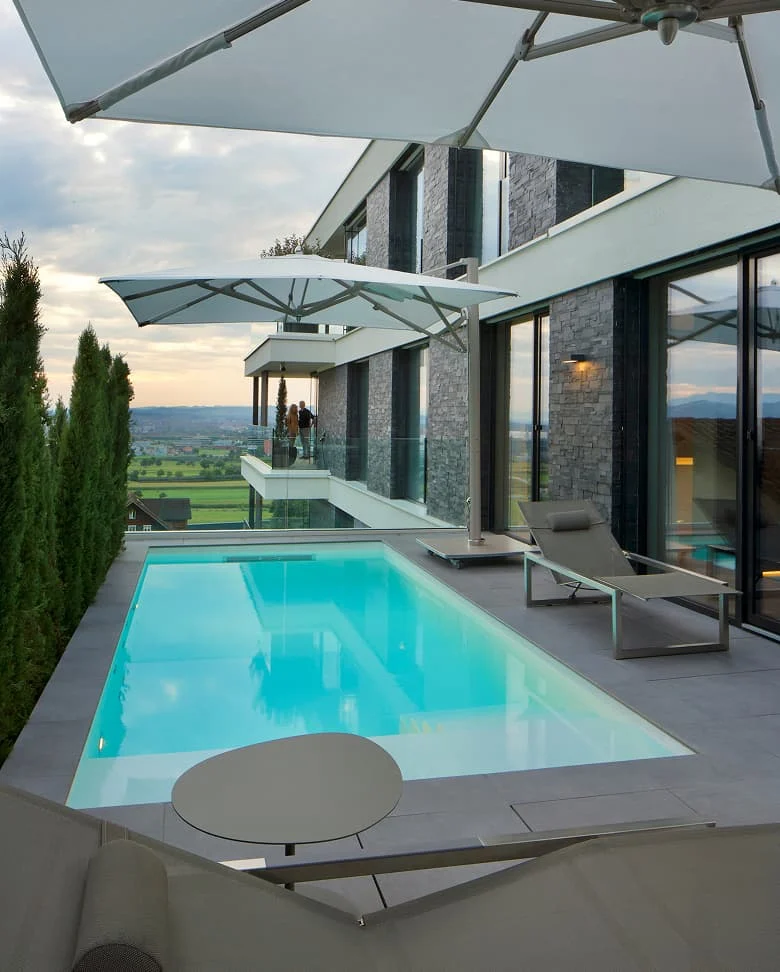 Pool auf einer Terrasse vor moderner Villa mit großen Glasflächen, Sonnenschirmen und Blick über eine Landschaft.