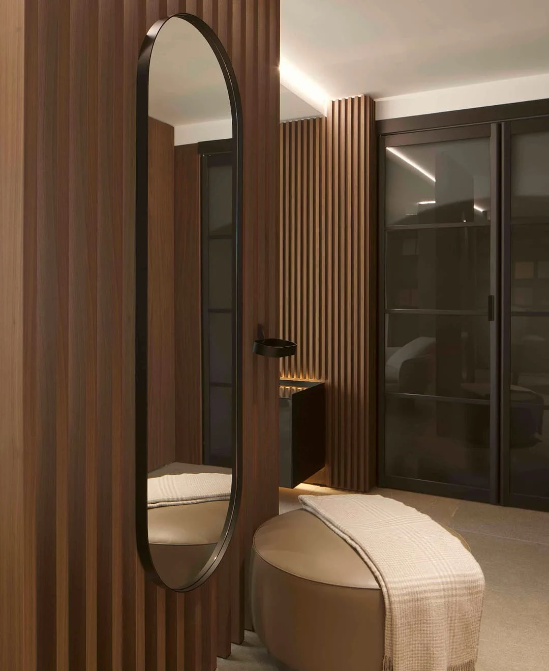 Moderner Ankleidebereich mit vertikalen Holzlamellen, elegantem ovalem Wandspiegel und rundem Pouf mit Decke im Trendline Showroom.