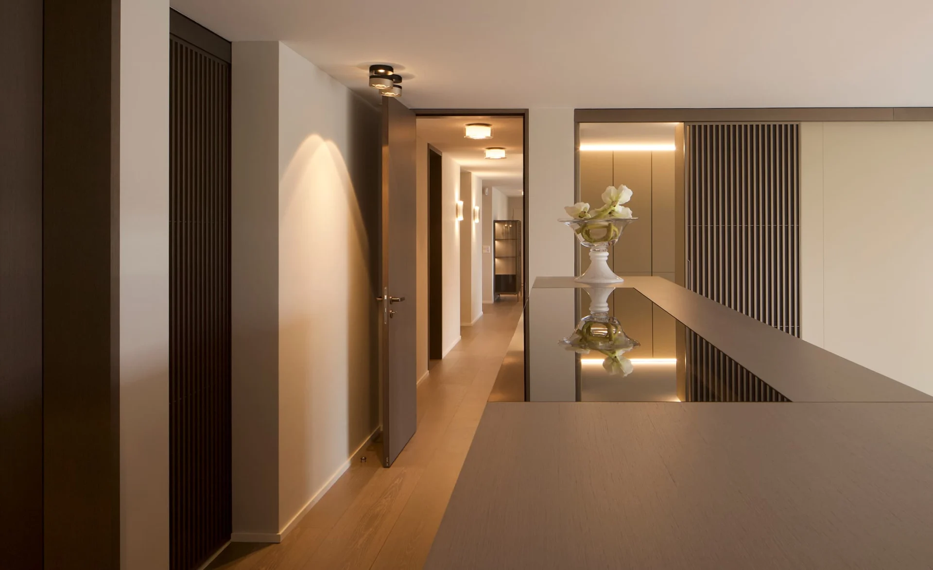 Moderner Flur mit warmen Holzelementen, offenen Türen und integrierter Wandbeleuchtung, im Vordergrund eine elegante Glasvase mit Blumen, die sich auf der glänzenden Oberfläche des Tisches spiegelt.
