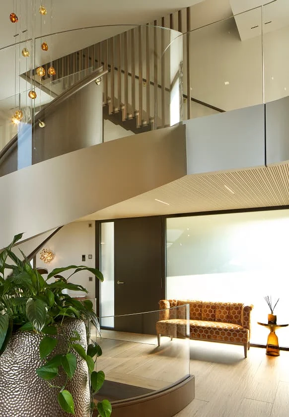 Moderner Innenraum mit geschwungener Treppe, Glasgeländer und Galerieebene über einem Sitzbereich.