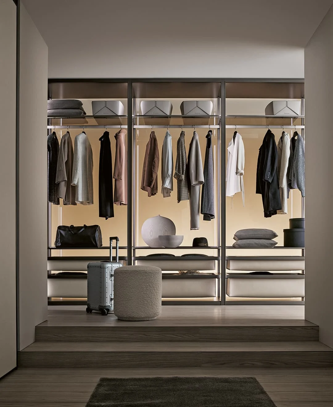 Offen gestalteter, moderner Kleiderschrank mit hängender Kleidung, Regalen, dekorativen Boxen und einem Hocker im Vordergrund.