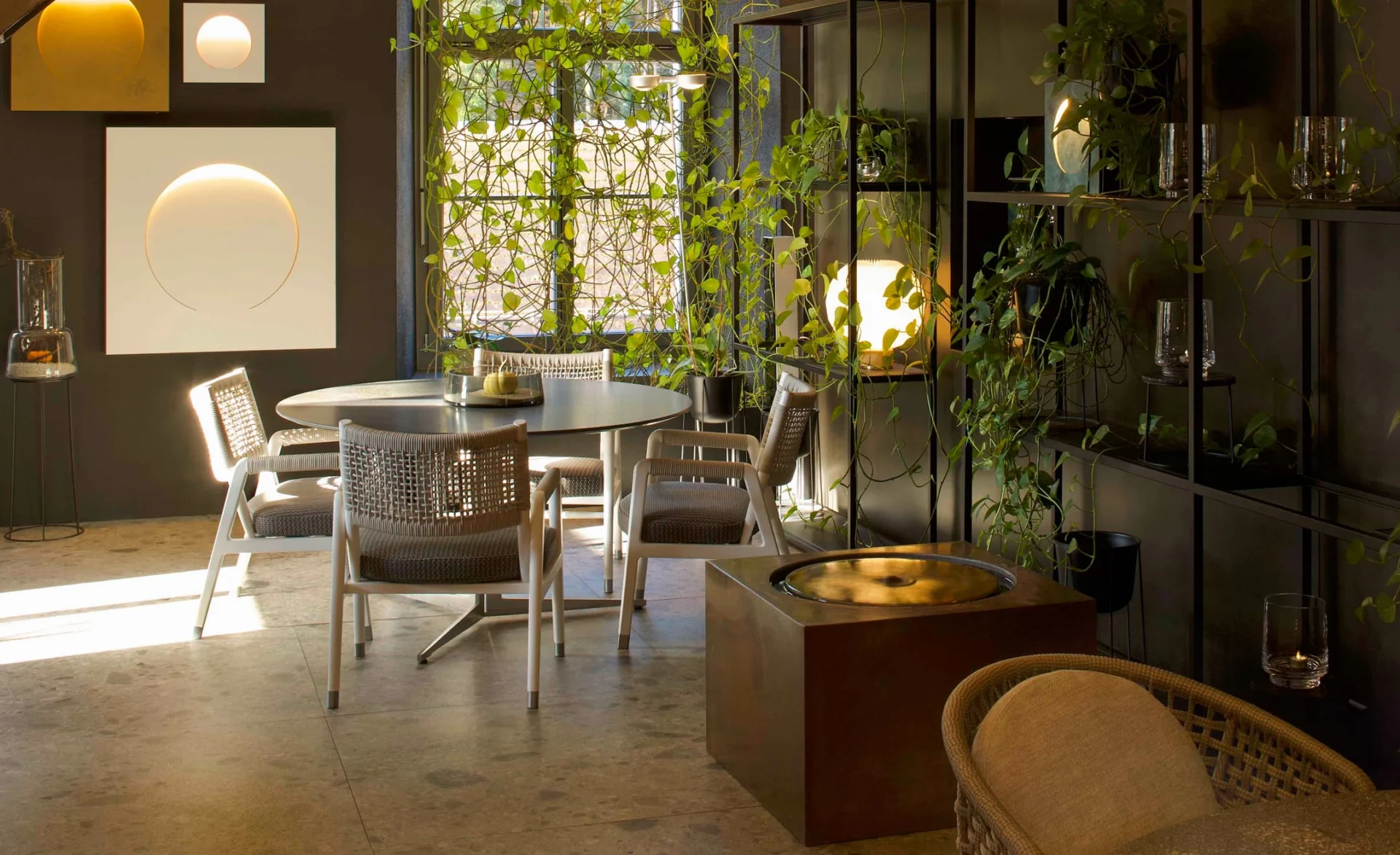 Moderner Essbereich mit runden Tisch, geflochtenen Stühlen und vielen hängenden Grünpflanzen, warm beleuchtet durch dekorative Wand- und Regalleuchten.