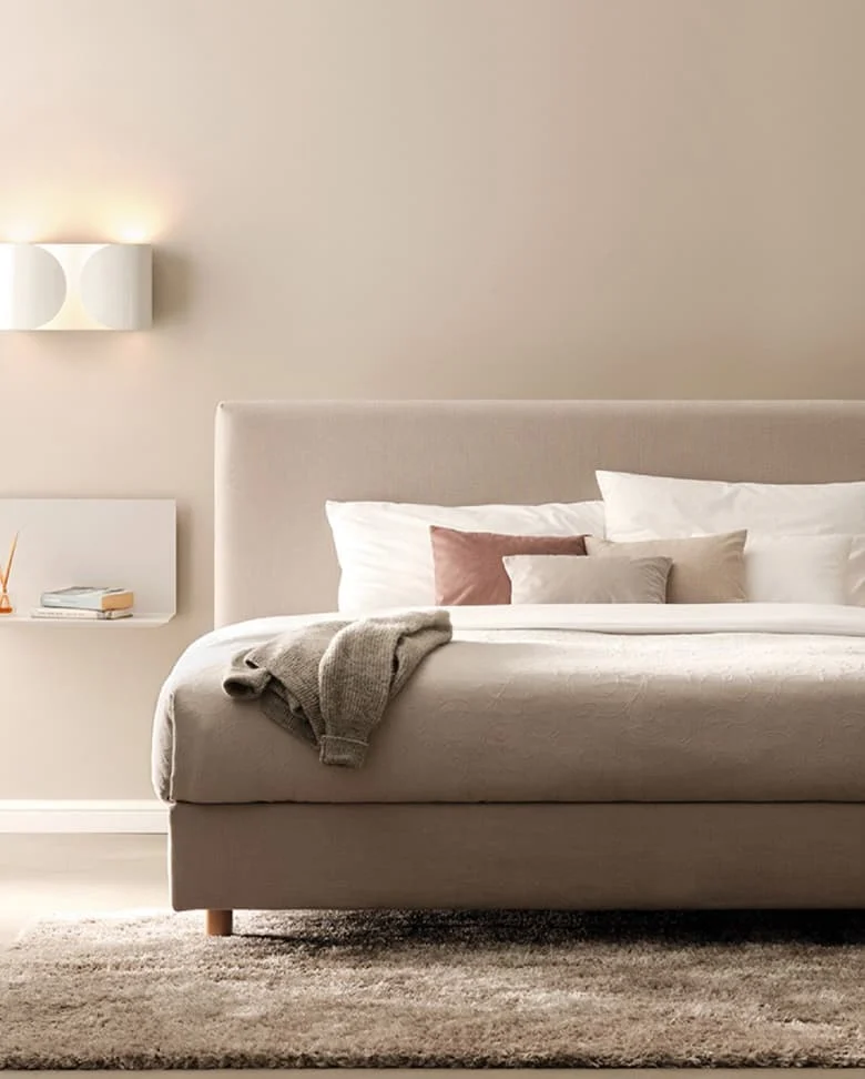 Modernes beige gepolstertes Bett mit weichen Kissen und Decke in einem ruhigen Schlafzimmer mit Wandlampe und hellem Teppich.