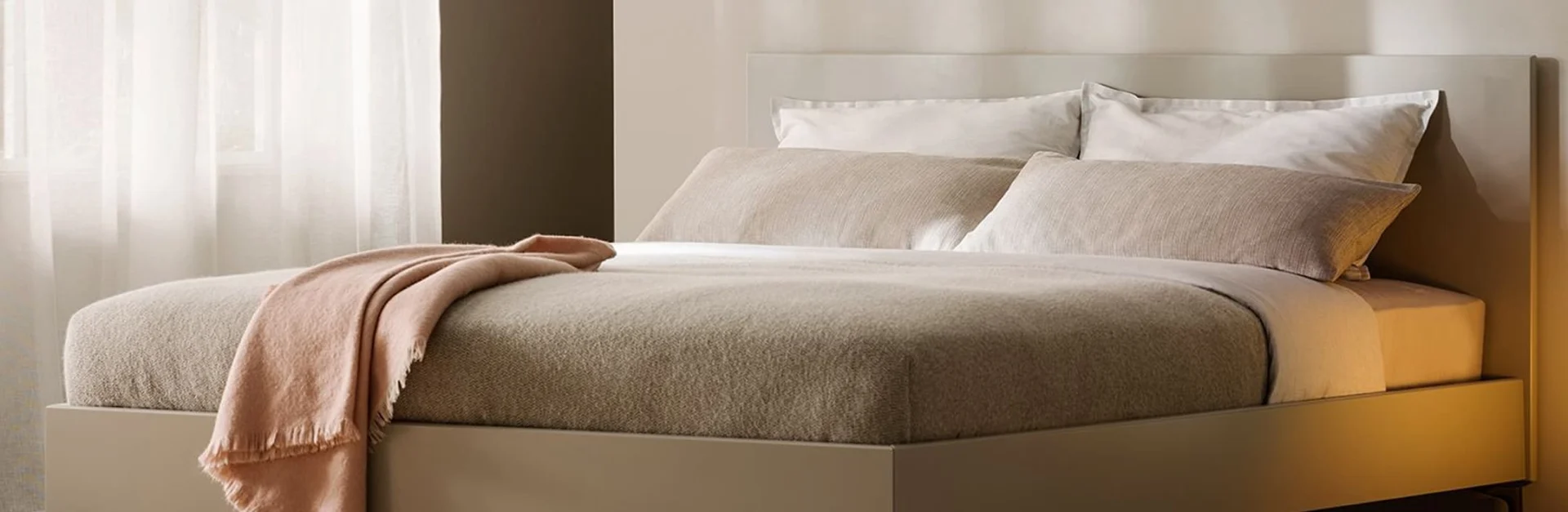 Modernes Bett mit beigen Textilien, weißen Kissen und einer roséfarbenen Decke in einem hellen, sanft beleuchteten Schlafzimmer.