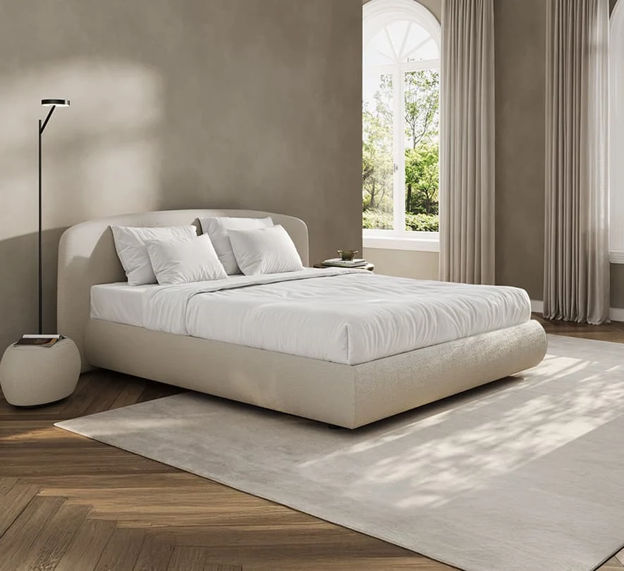 Modernes cremefarbenes Polsterbett mit abgerundetem Kopfteil in einem eleganten Schlafzimmer mit großen Fenstern, hellen Vorhängen und warmem Holzboden.