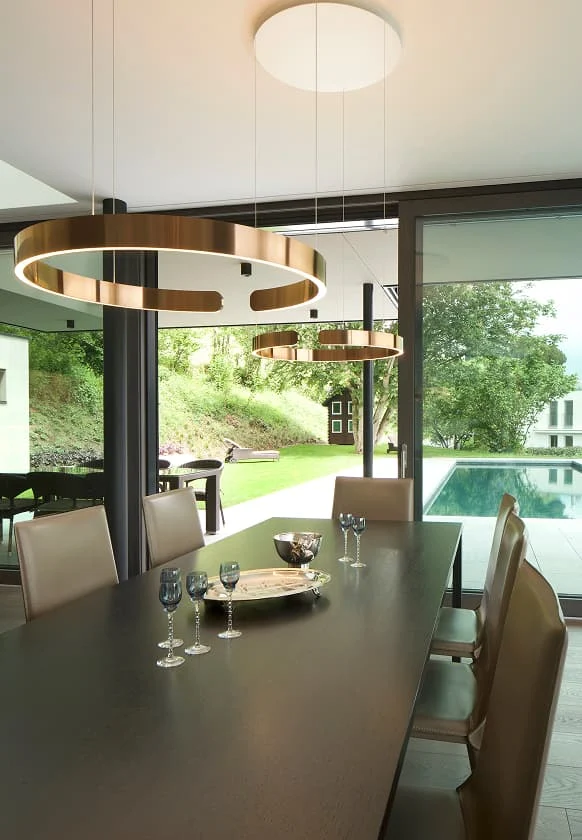 Modernes Esszimmer mit großem dunklen Esstisch, eleganten Lederstühlen, goldenen Ringpendelleuchten und Blick auf Garten und Pool.