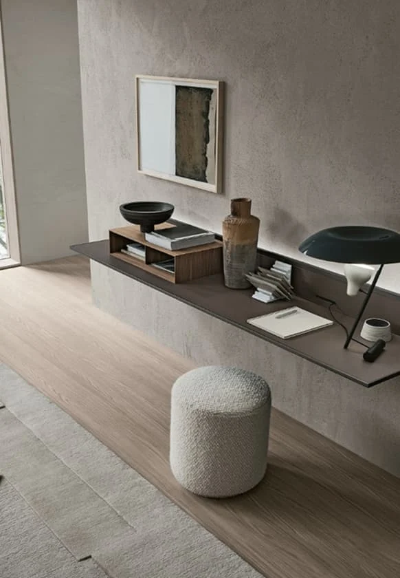 Modernes Interieur mit schwebendem Wandregal, Dekoobjekten, Büchern und einer Designer-Tischlampe, ergänzt durch einen hellen Hocker auf Holzfußboden.