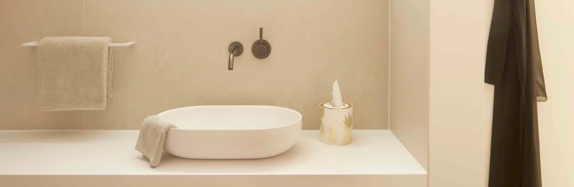 Modernes, minimalistisch gestaltetes Badezimmer mit weißer Waschtischschale, Wandarmatur, beigem Handtuch und dekorativer Papiertuchdose.