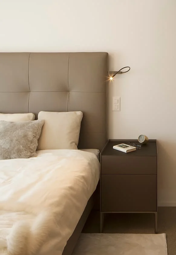 Detailansicht eines modernen Schlafzimmers mit gepolstertem Kopfteil, hellen Kissen und braunem Nachttisch.