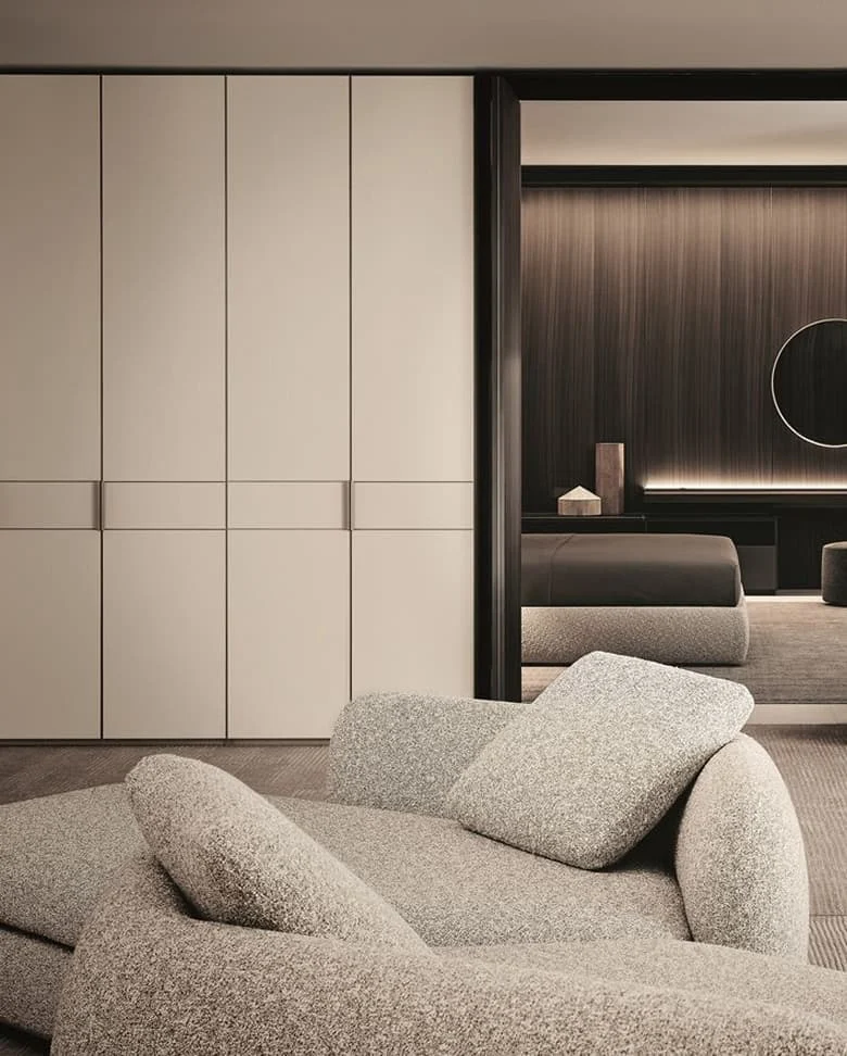 Modernes Schlafzimmer mit hellen Einbauschränken, gemütlicher beigefarbener Lounge-Liege im Vordergrund und Blick in einen zweiten Raum mit dunkler Holzverkleidung und rundem Spiegel.